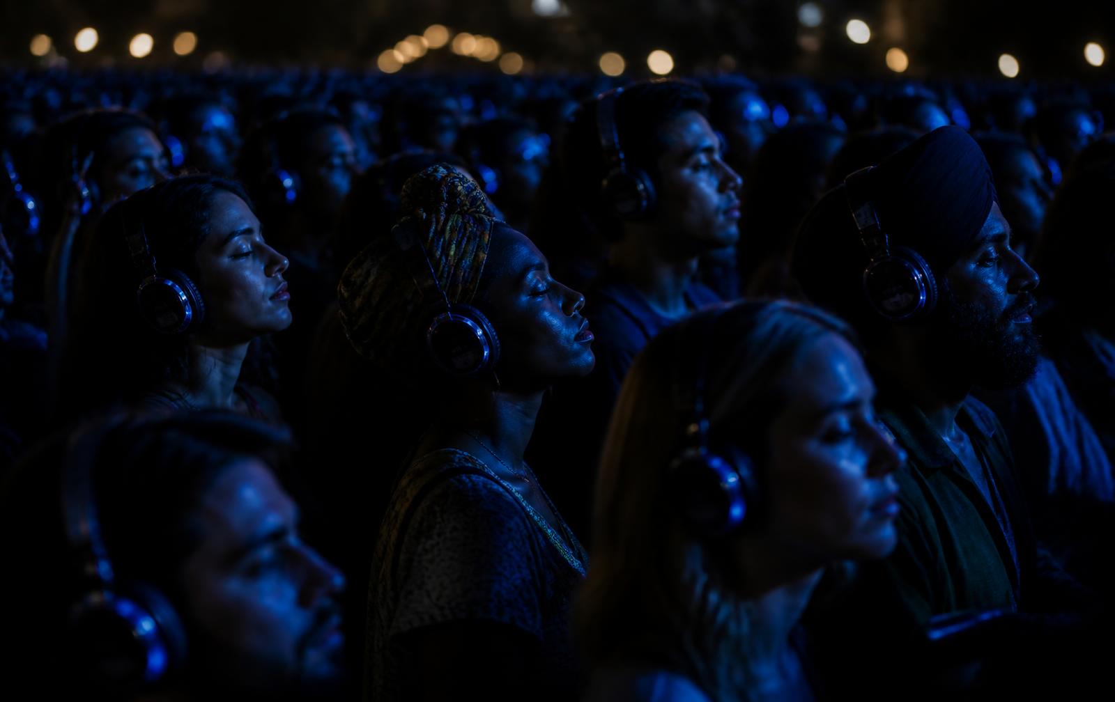 A diverse international crowd listening on headphones, lit in deep blue